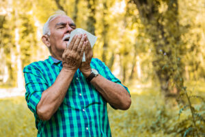Man with gray hair standing in a wooded area, holding a tissue in his hands and preparing to sneeze